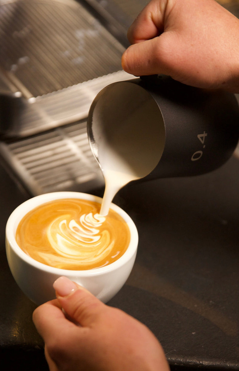 Barista pouring milk into a cup of coffee on a dark counter.